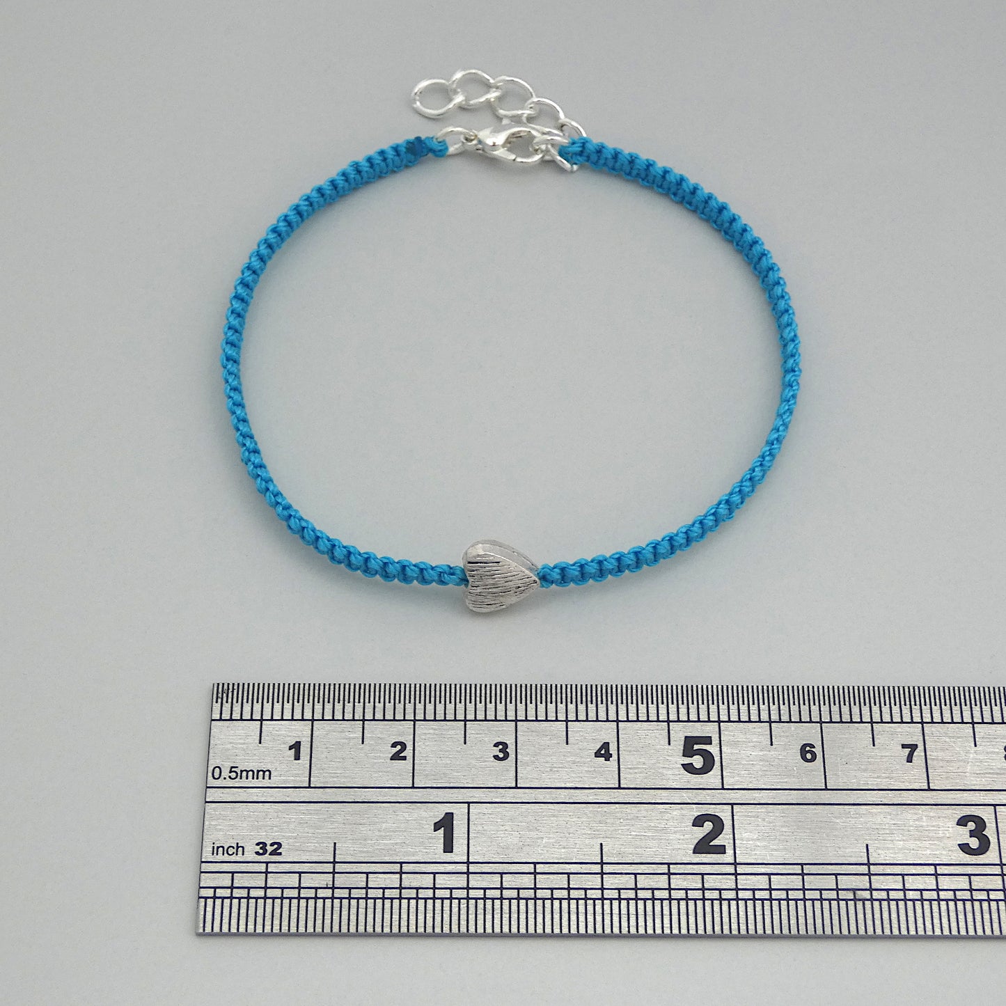 Blue seven inch macramé bracelet with silver plated textured heart charm, lobster clasp and one inch extender chain next to a ruler.