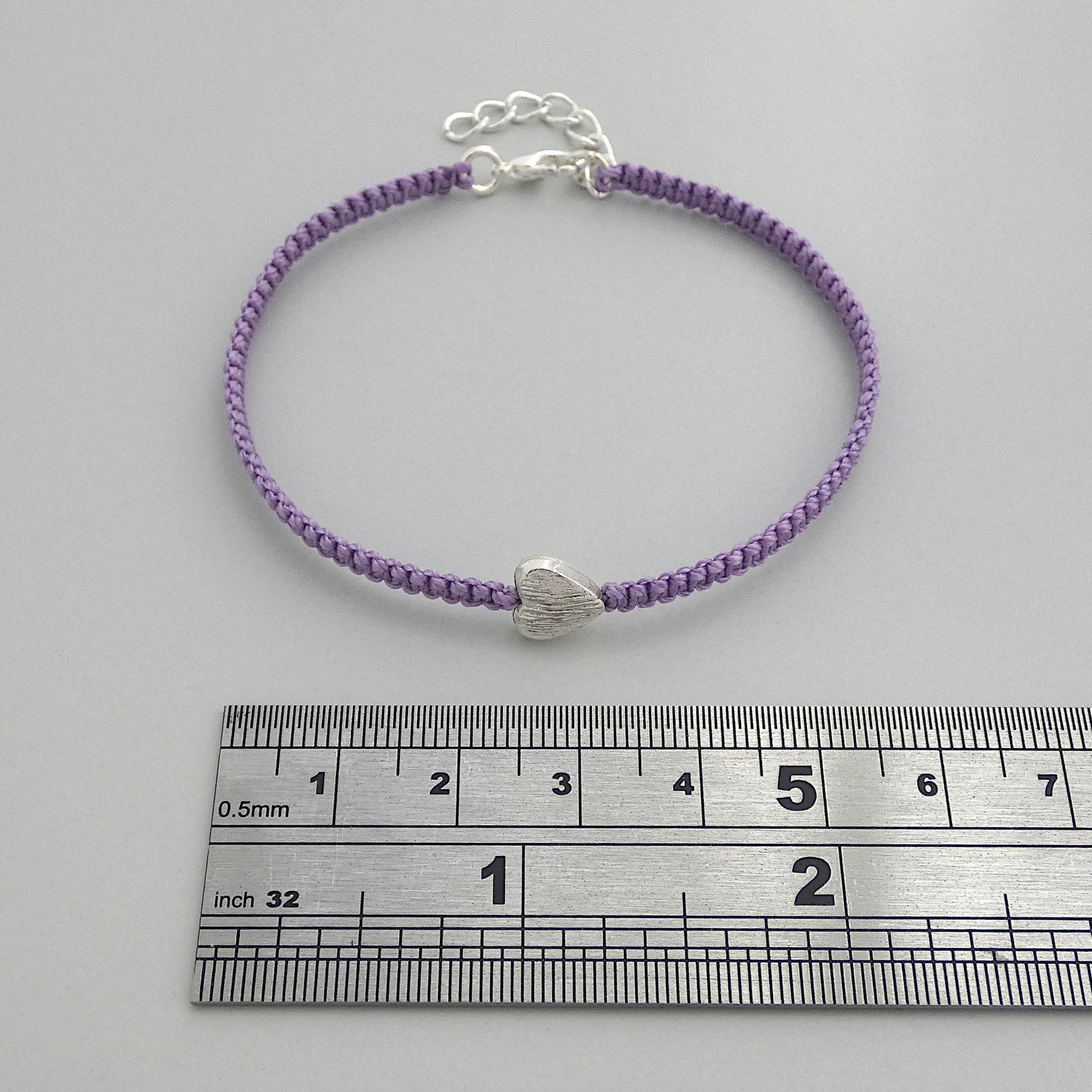 Orchid purple seven inch macramé bracelet with silver plated textured heart charm, lobster clasp and one inch extender chain next to a ruler.