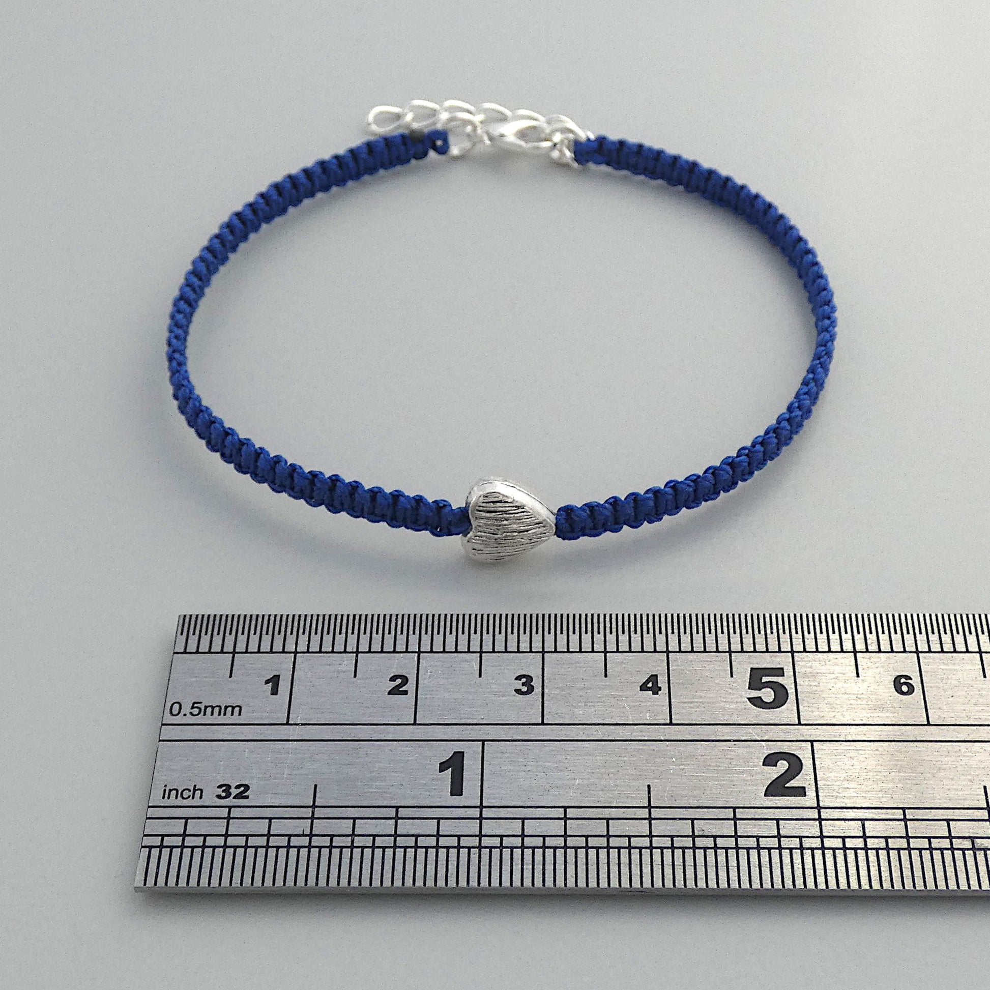 Royal blue seven inch macramé bracelet with silver plated textured heart charm, lobster clasp and one inch extender chain next to a ruler.