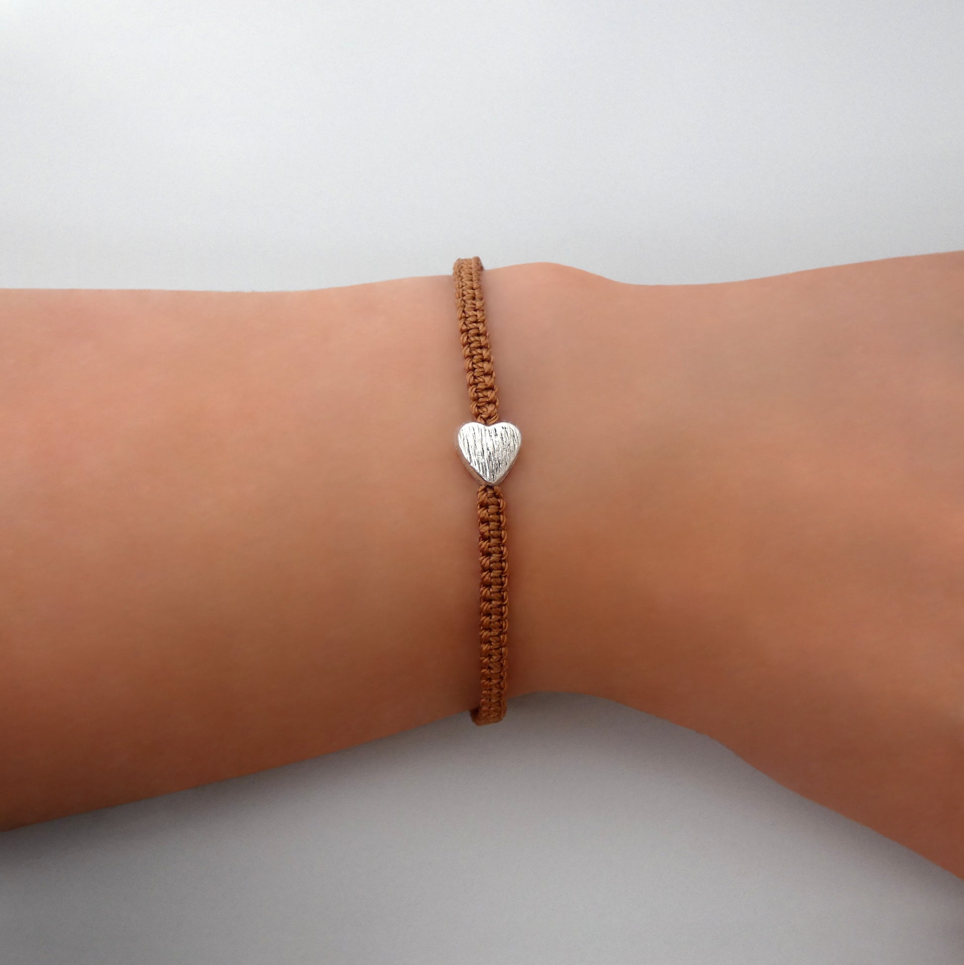 Copper coloured seven inch macramé bracelet with silver plated textured heart charm, lobster clasp and one inch extender chain on a woman's wrist.