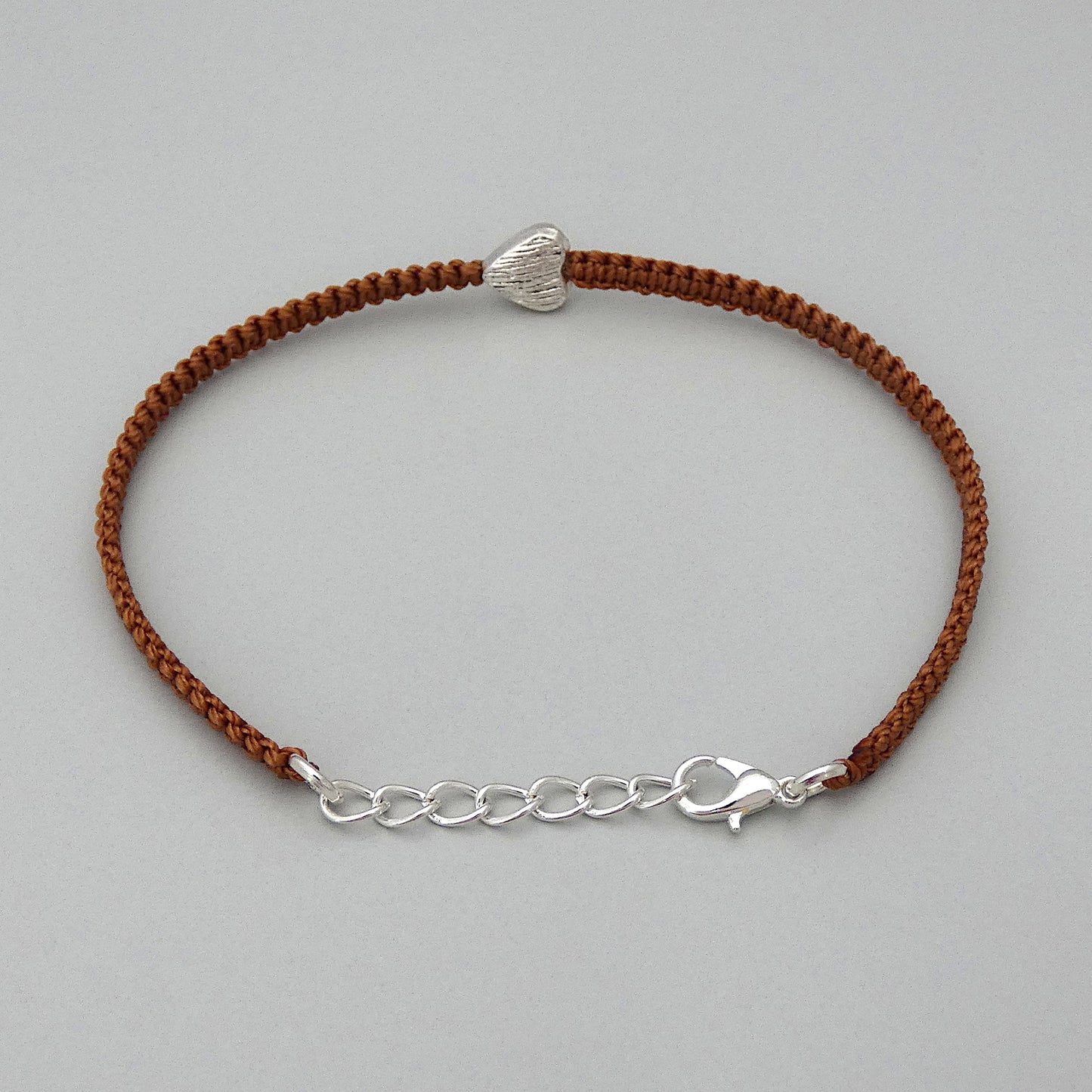 Silver plated lobster claw clasp attached to the end of a one inch extender chain on a copper coloured seven inch macramé bracelet with silver plated textured heart charm.