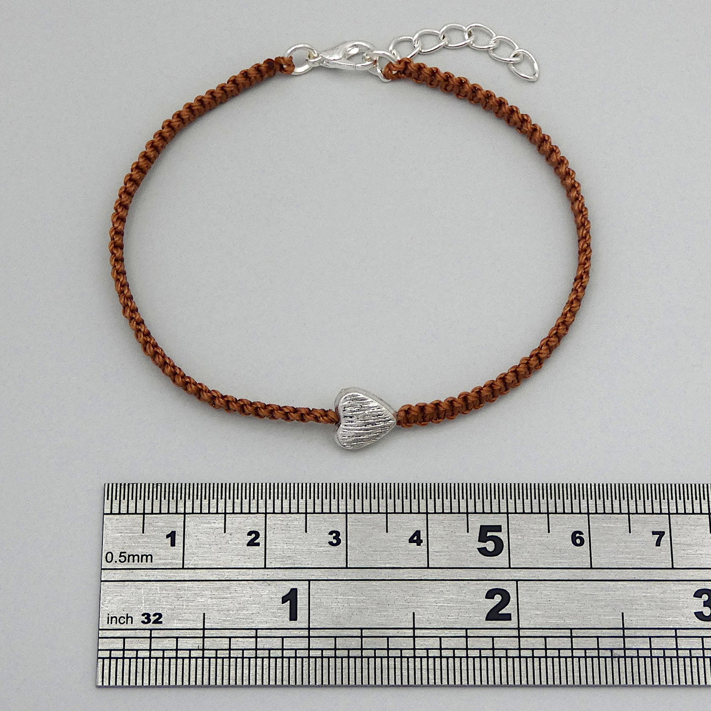 Copper coloured seven inch macramé bracelet with silver plated textured heart charm, lobster clasp and one inch extender chain next to a ruler.