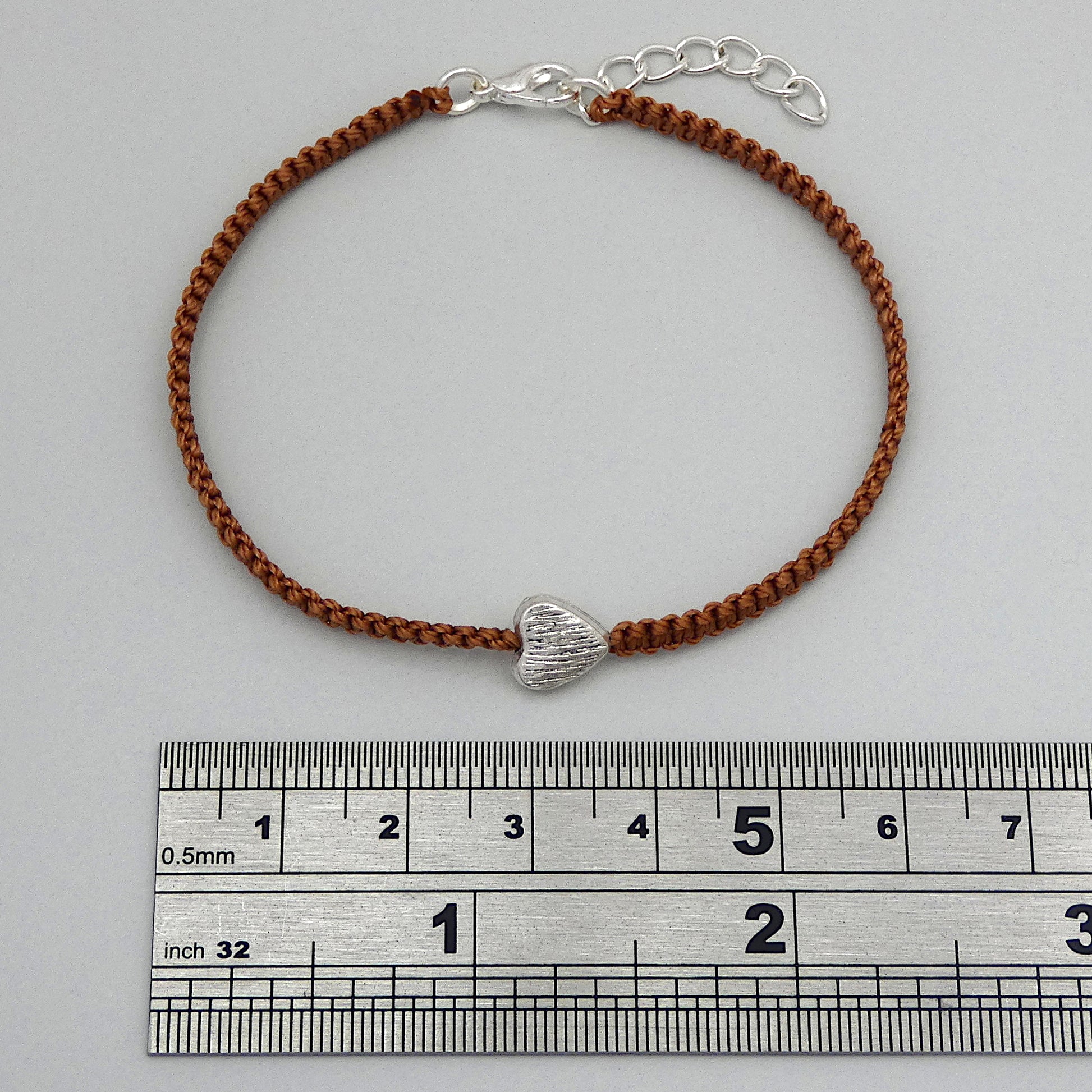 Copper coloured seven inch macramé bracelet with silver plated textured heart charm, lobster clasp and one inch extender chain next to a ruler.
