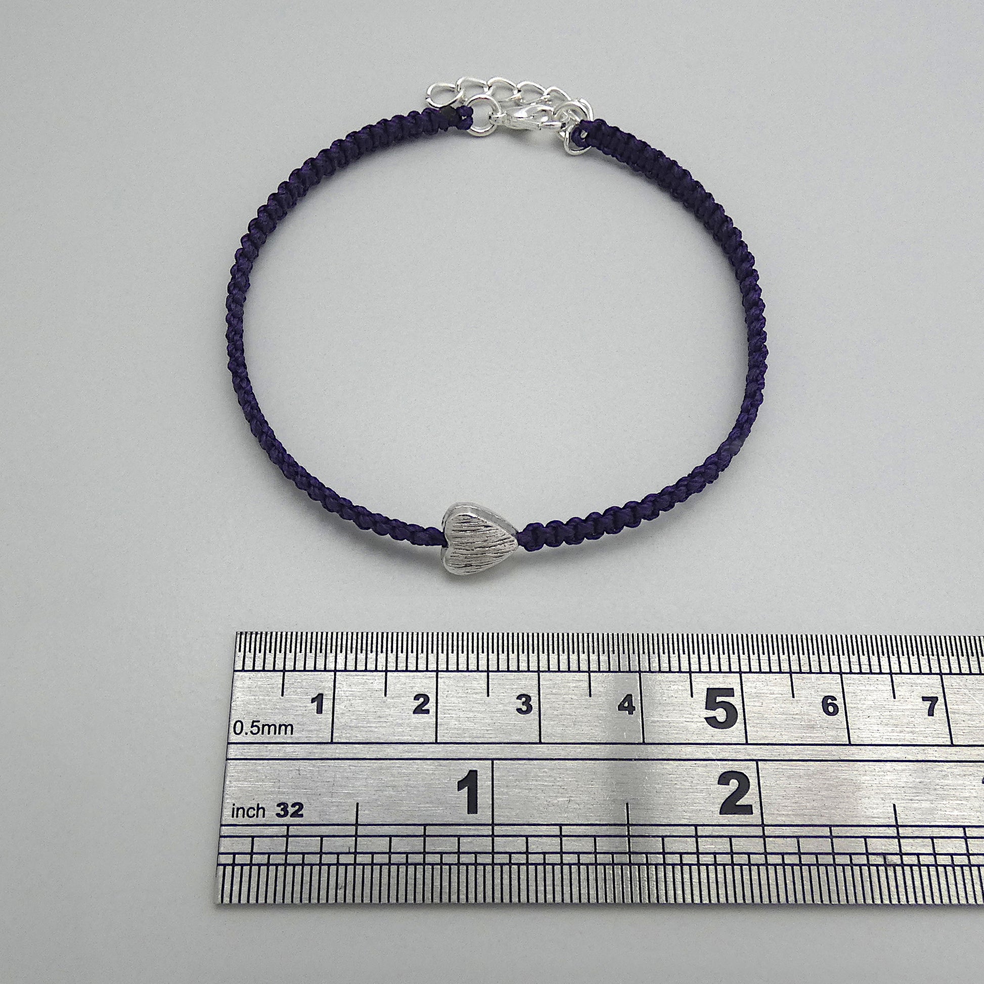 Dark purple seven inch macramé bracelet with silver plated textured heart charm, lobster clasp and one inch extender chain next to a ruler.