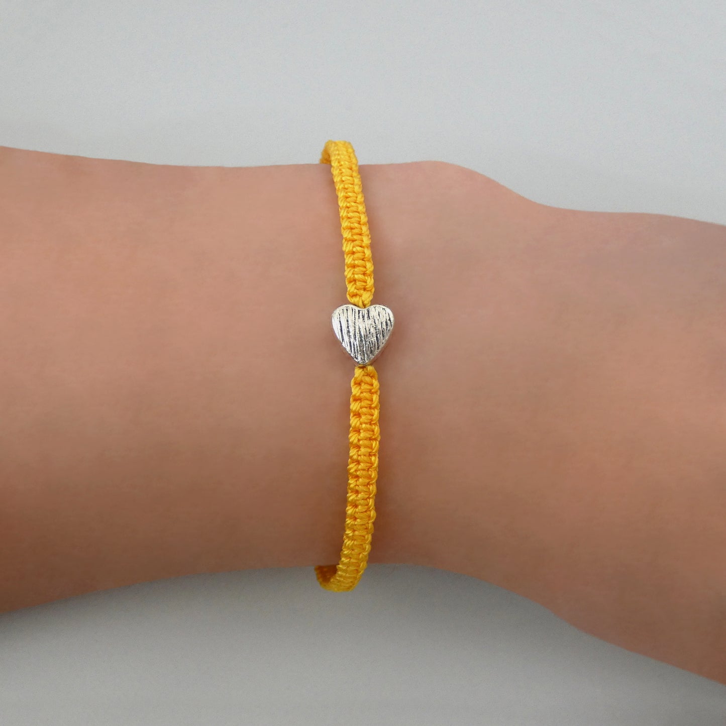 Golden yellow seven inch macramé bracelet with silver plated textured heart charm, lobster clasp and one inch extender chain on a woman's wrist.