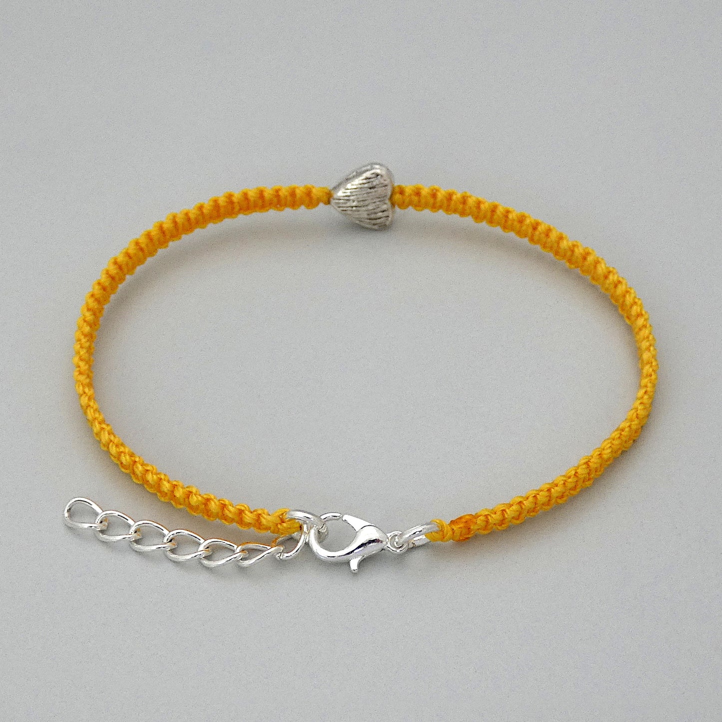 Silver plated lobster claw clasp and one inch extender chain on a golden yellow seven inch macramé bracelet with silver plated textured heart charm.