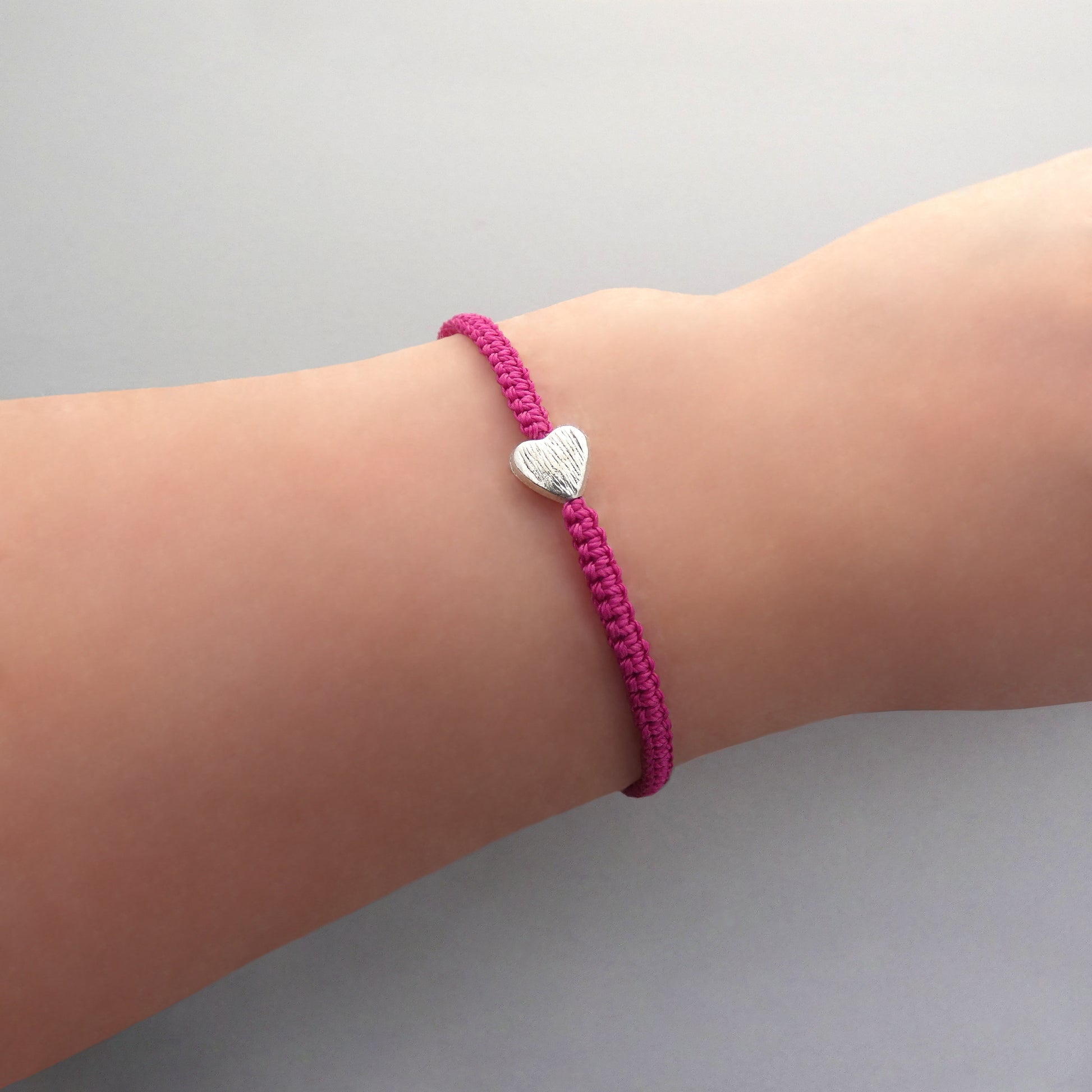 Magenta seven inch macramé bracelet with silver plated textured heart charm, lobster clasp and one inch extender chain on a woman's wrist.
