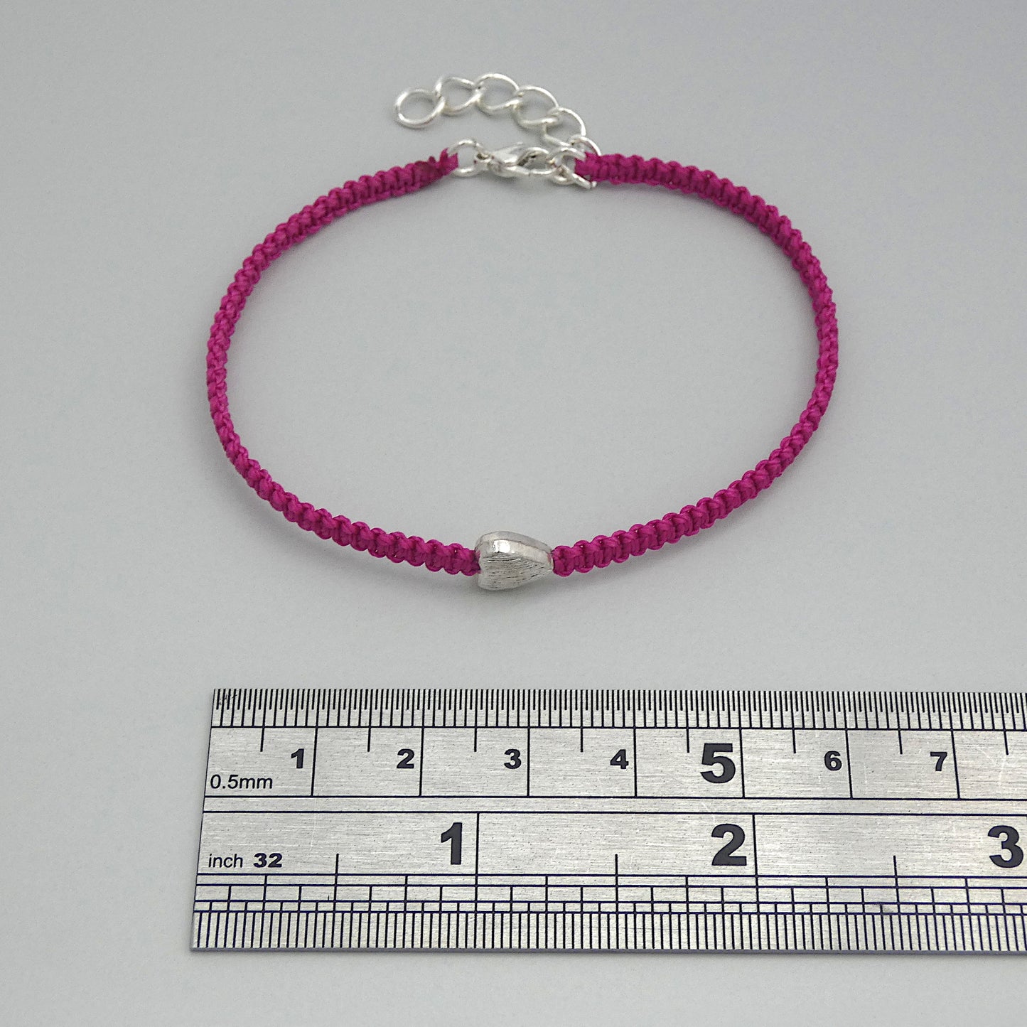 Magenta seven inch macramé bracelet with silver plated textured heart charm, lobster clasp and one inch extender chain next to a ruler.
