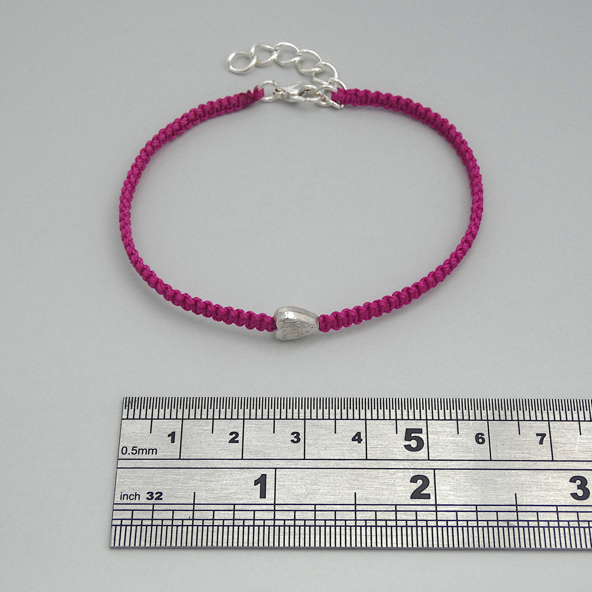 Magenta seven inch macramé bracelet with silver plated textured heart charm, lobster clasp and one inch extender chain next to a ruler.
