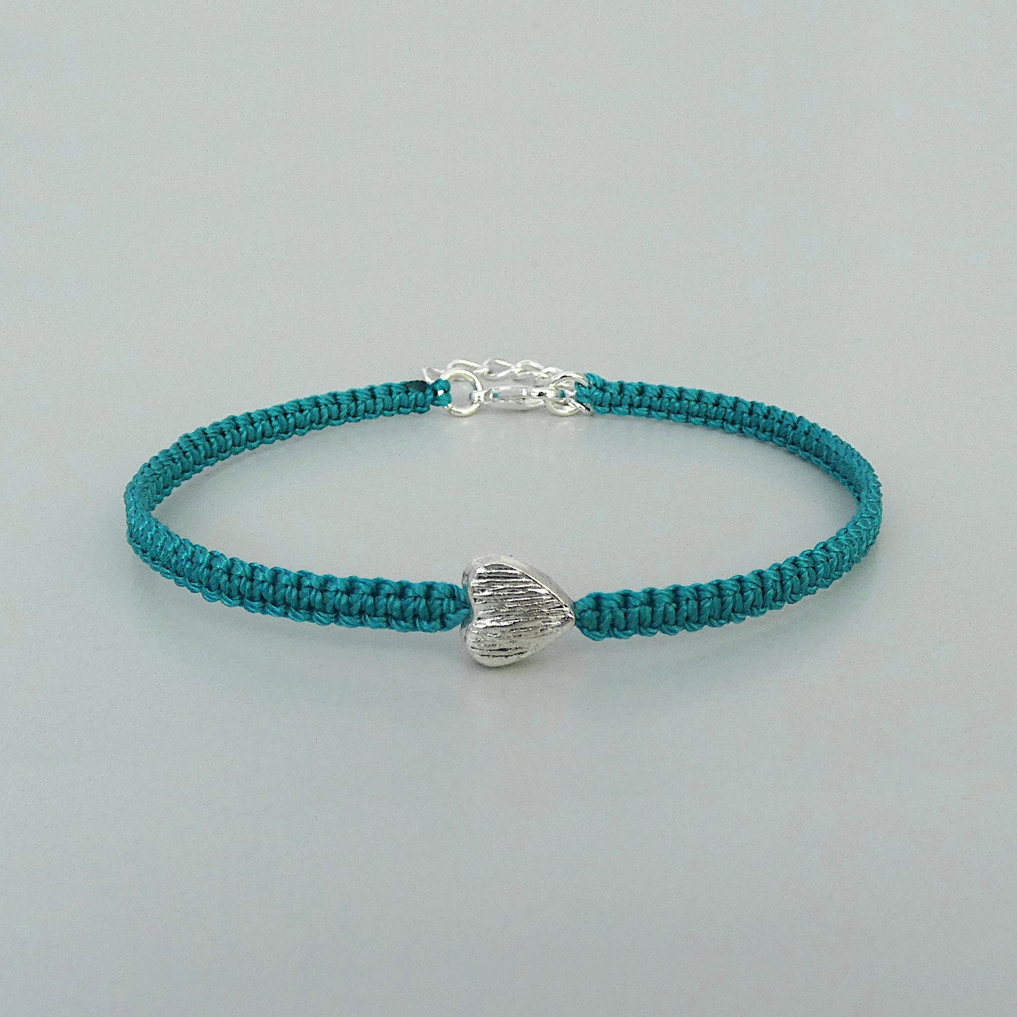 Teal green seven inch macramé bracelet with silver plated textured heart charm, lobster clasp and one inch extender chain.