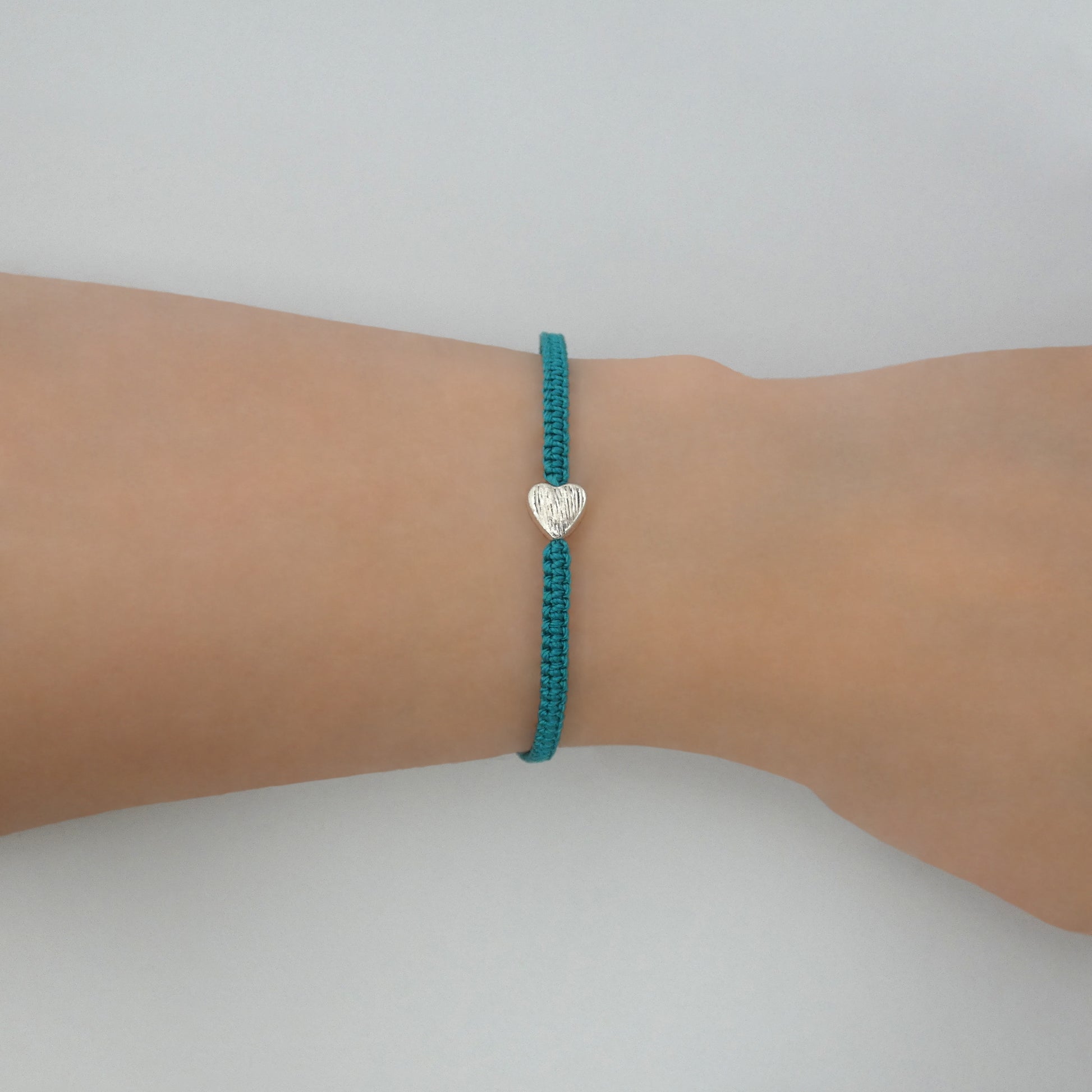 Teal green seven inch macramé bracelet with silver plated textured heart charm, lobster clasp and one inch extender chain on a women's wrist.