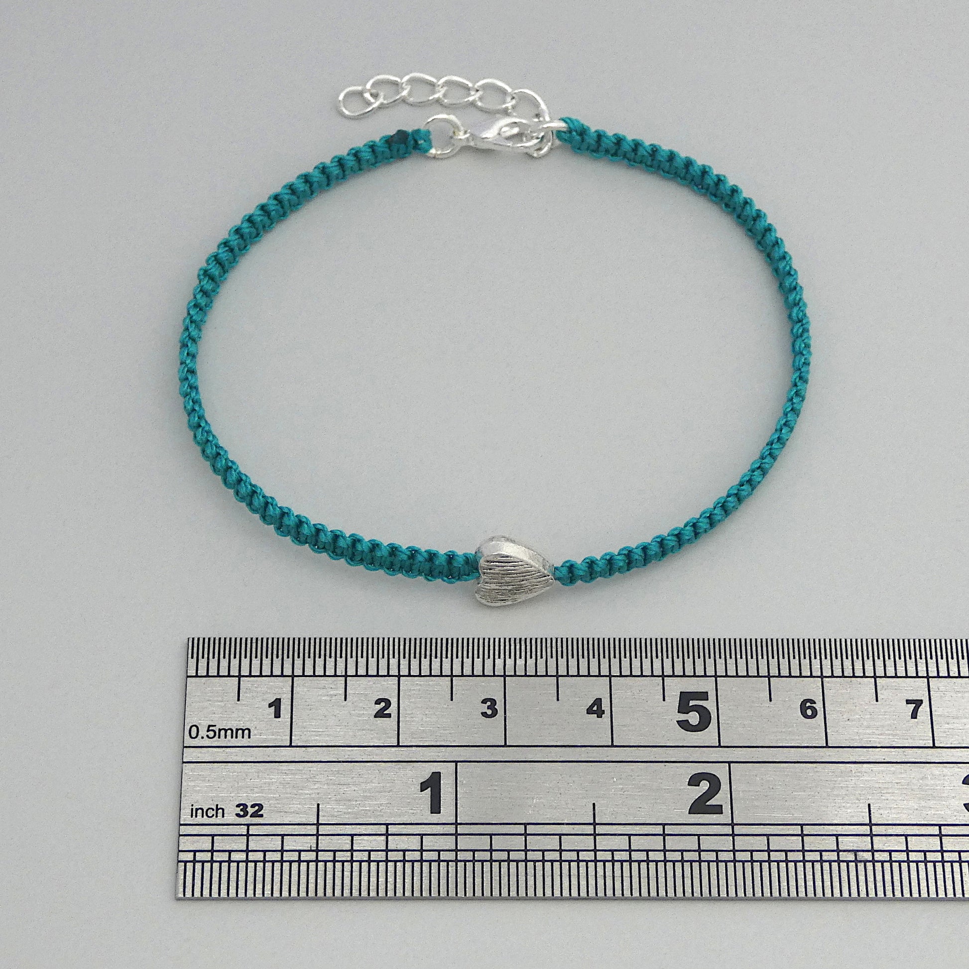 Teal green seven inch macramé bracelet with silver plated textured heart charm, lobster clasp and one inch extender chain next to a ruler.