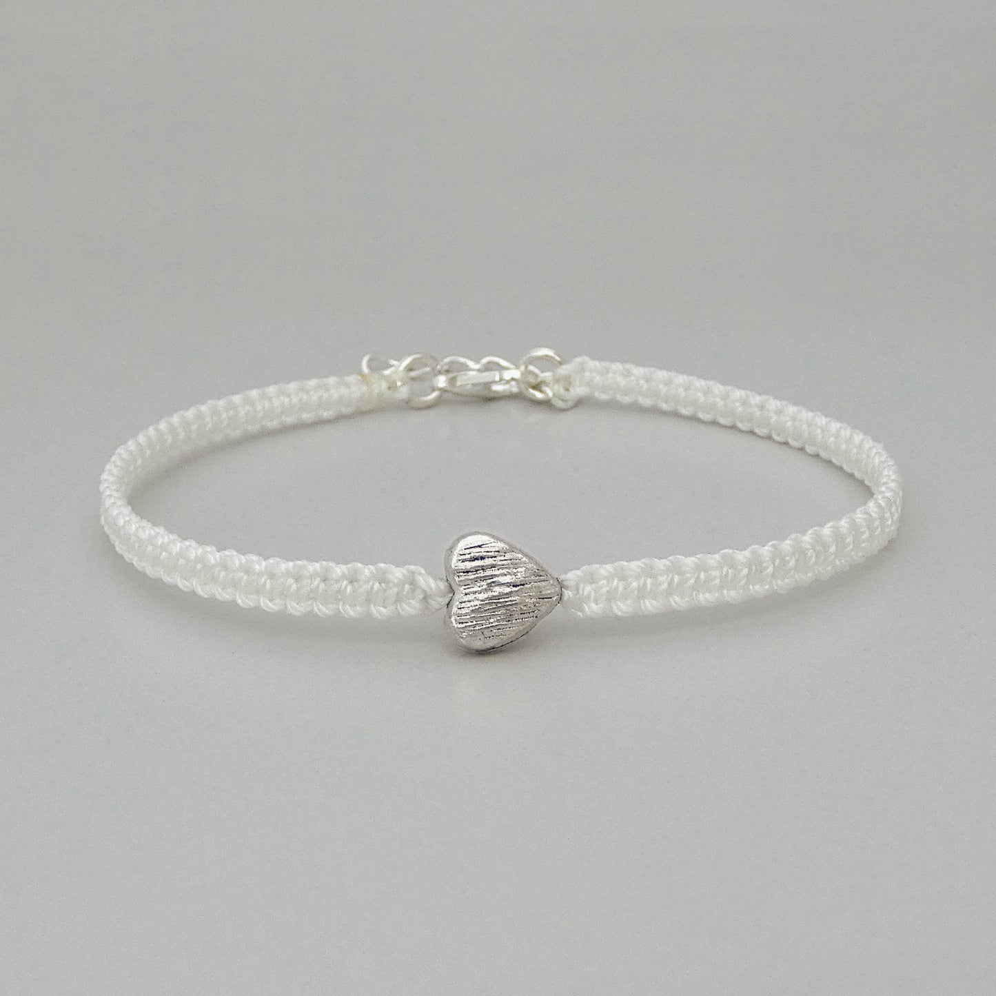 White seven inch macramé bracelet with silver plated textured heart charm, lobster clasp and one inch extender chain.