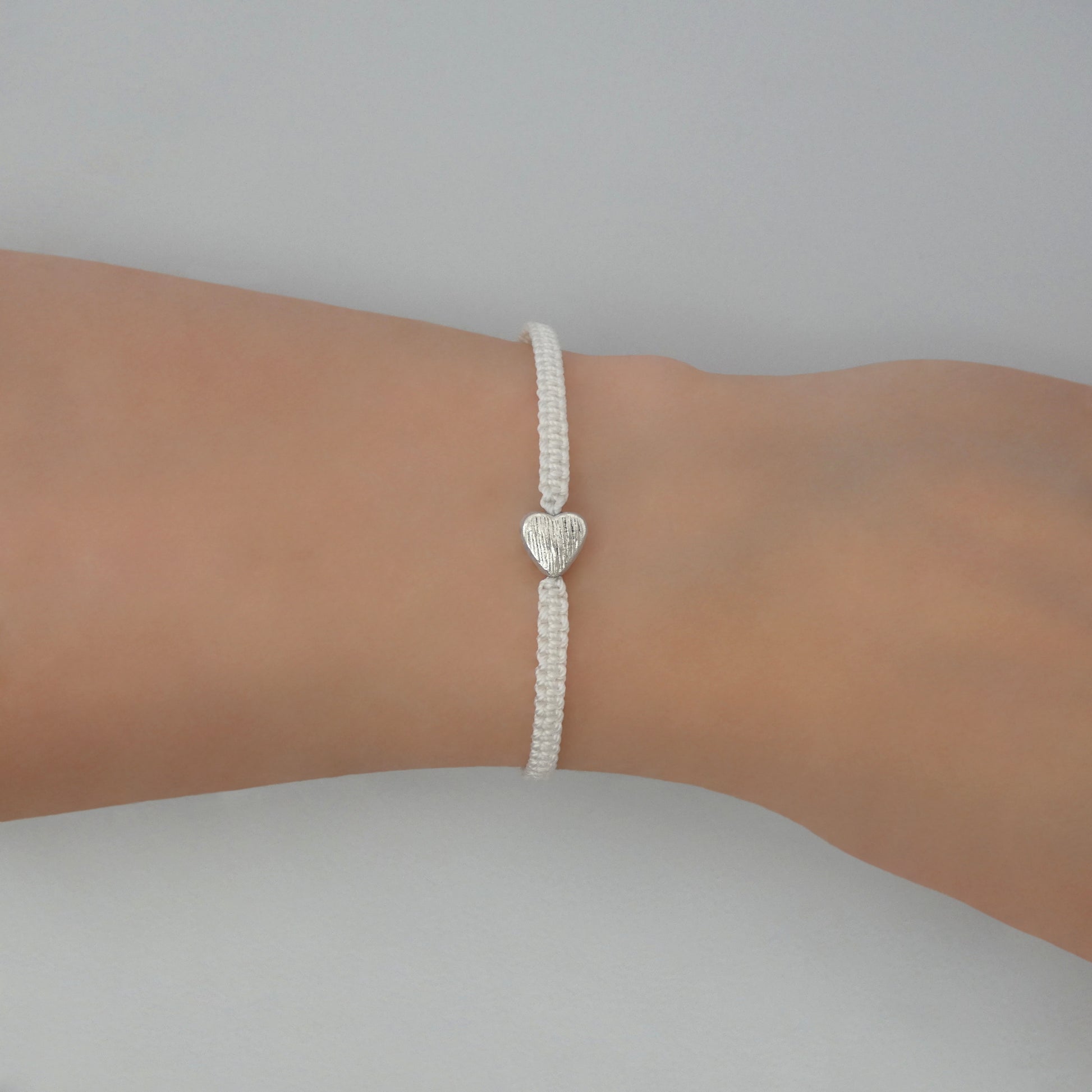 White seven inch macramé bracelet with silver plated textured heart charm, lobster clasp and one inch extender chain on a woman's wrist.