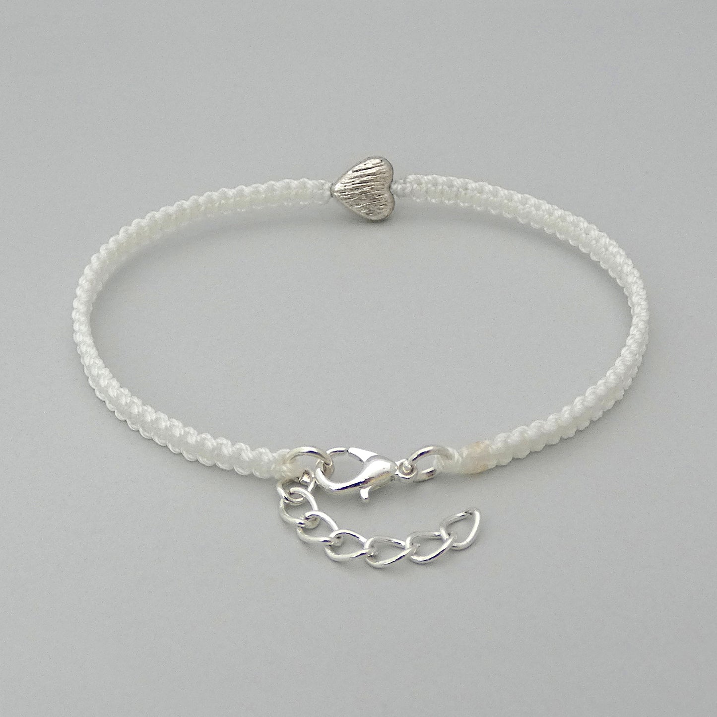 Silver plated lobster claw clasp and one inch extender chain on a white seven inch macramé bracelet with silver plated textured heart charm.