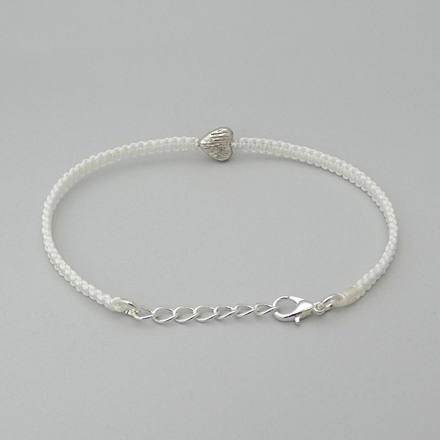 Silver plated lobster claw clasp attached to the end of a one inch extender chain on a white seven inch macramé bracelet with silver plated textured heart charm.