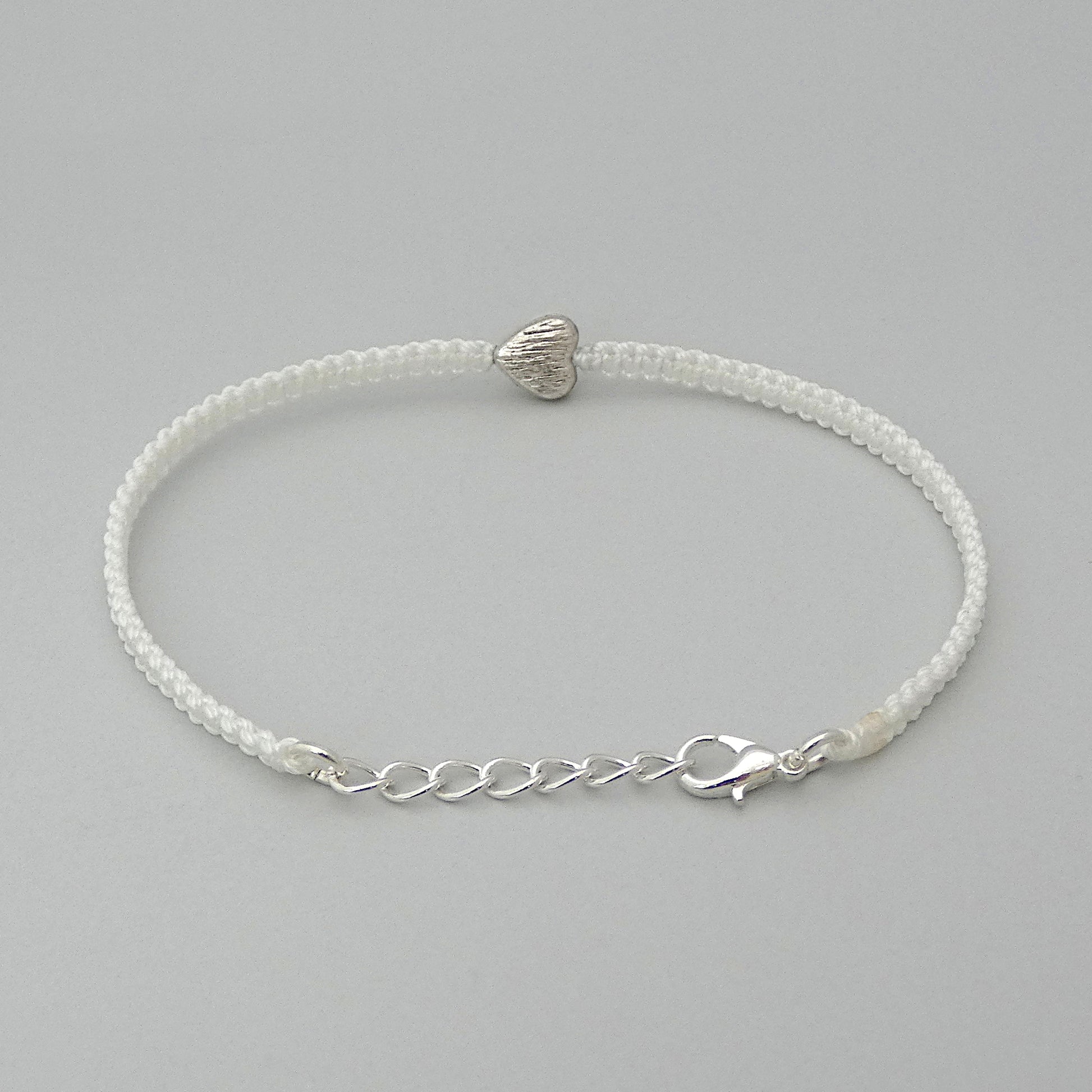 Silver plated lobster claw clasp attached to the end of a one inch extender chain on a white seven inch macramé bracelet with silver plated textured heart charm.