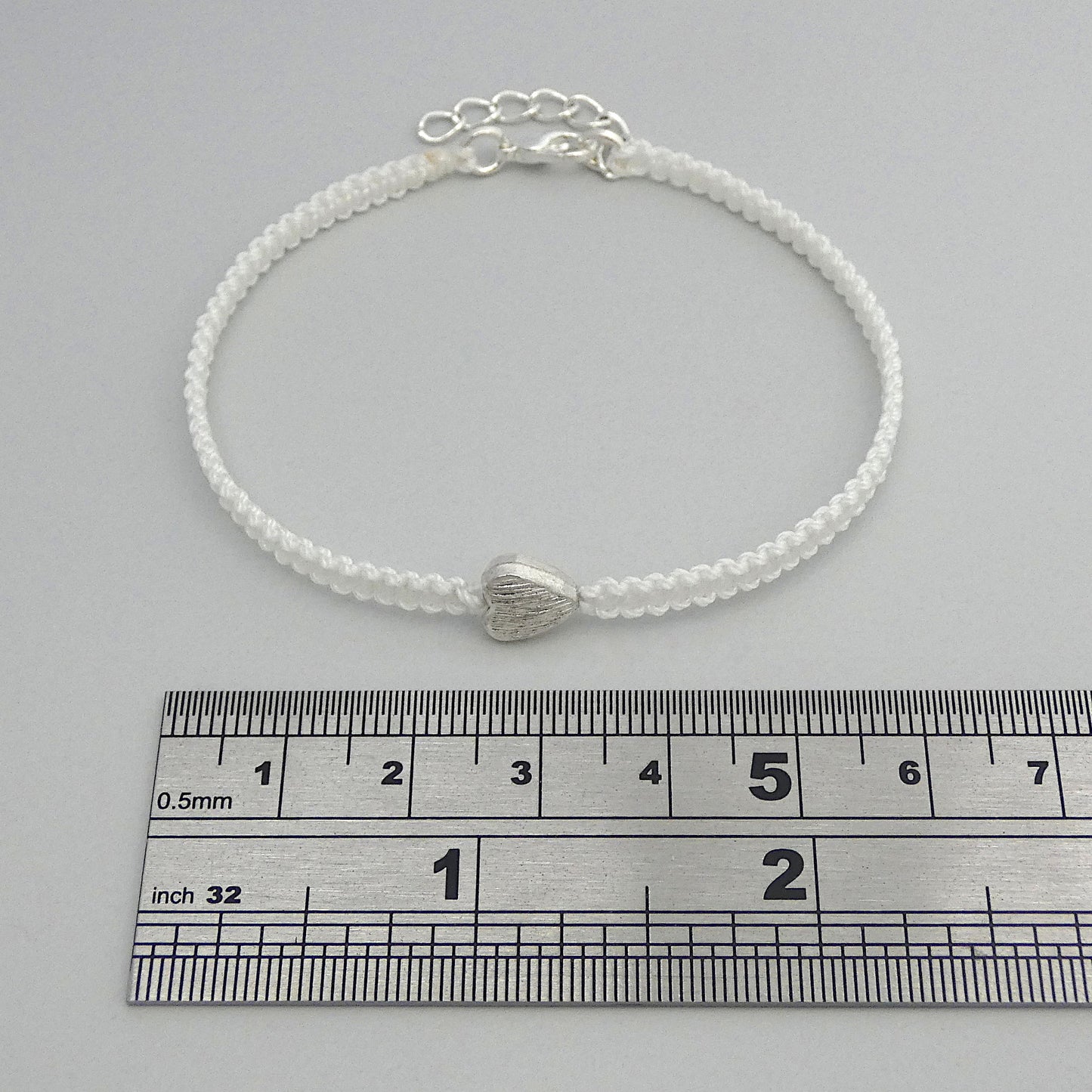 White seven inch macramé bracelet with silver plated textured heart charm, lobster clasp and one inch extender chain next to a ruler.