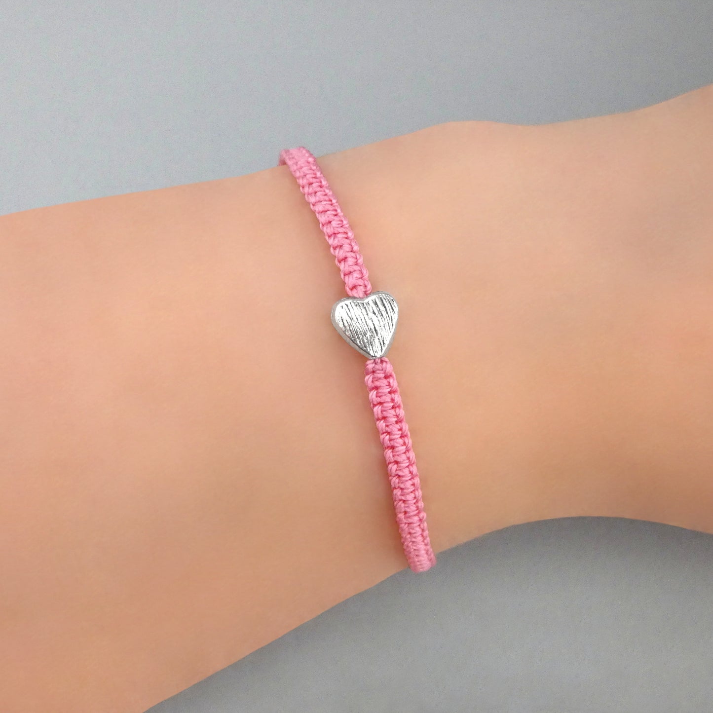 Pink seven inch macramé bracelet with silver plated textured heart charm, lobster clasp and one inch extender chain on a woman's wrist.