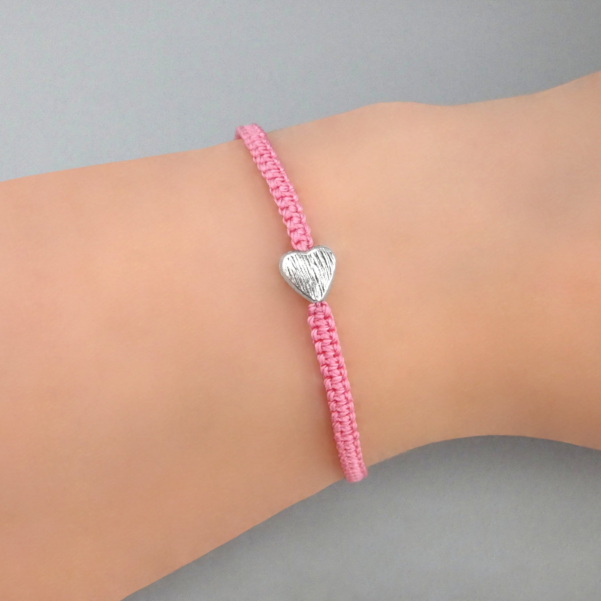 Pink seven inch macramé bracelet with silver plated textured heart charm, lobster clasp and one inch extender chain on a woman's wrist.