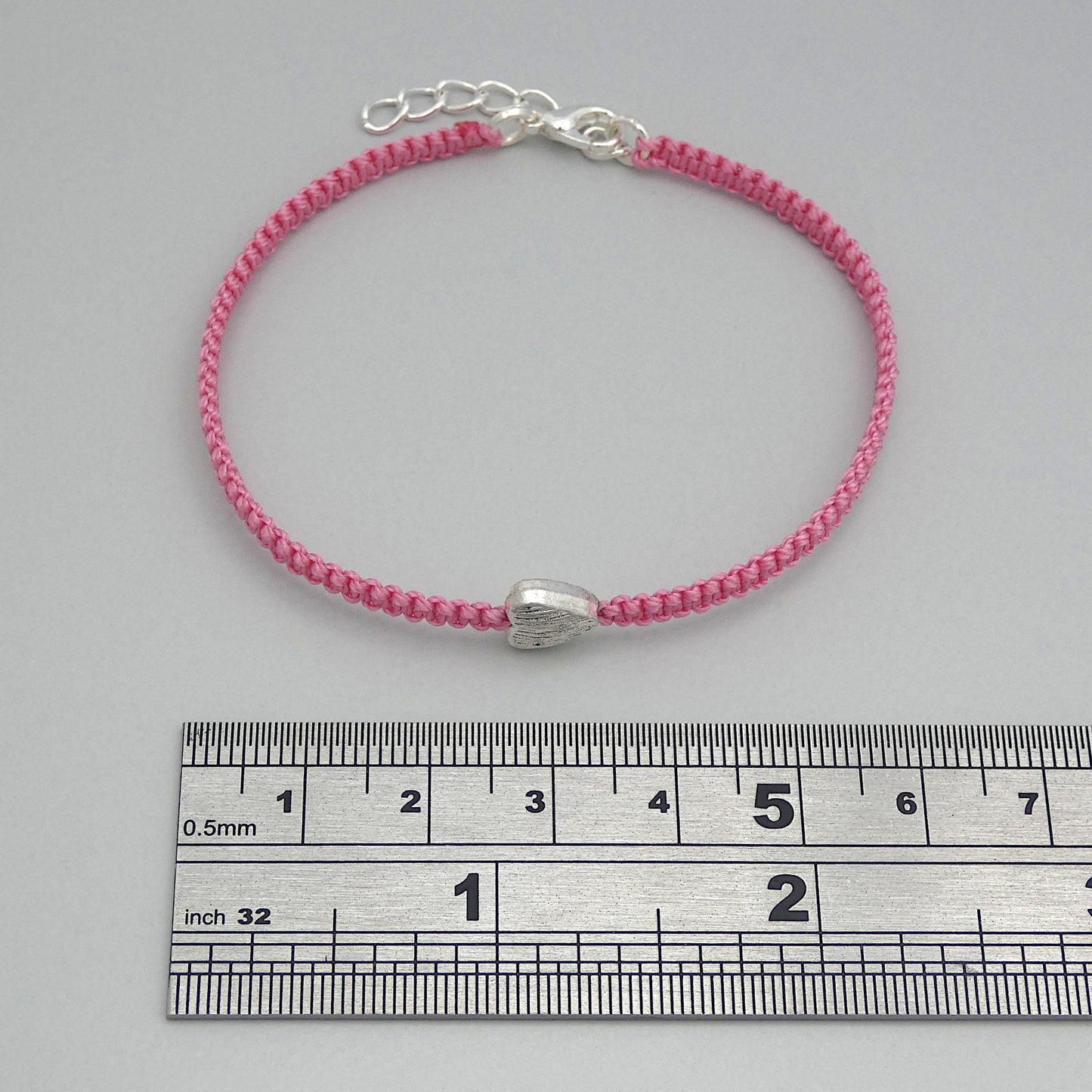 Pink seven inch macramé bracelet with silver plated textured heart charm, lobster clasp and one inch extender chain next to a ruler.