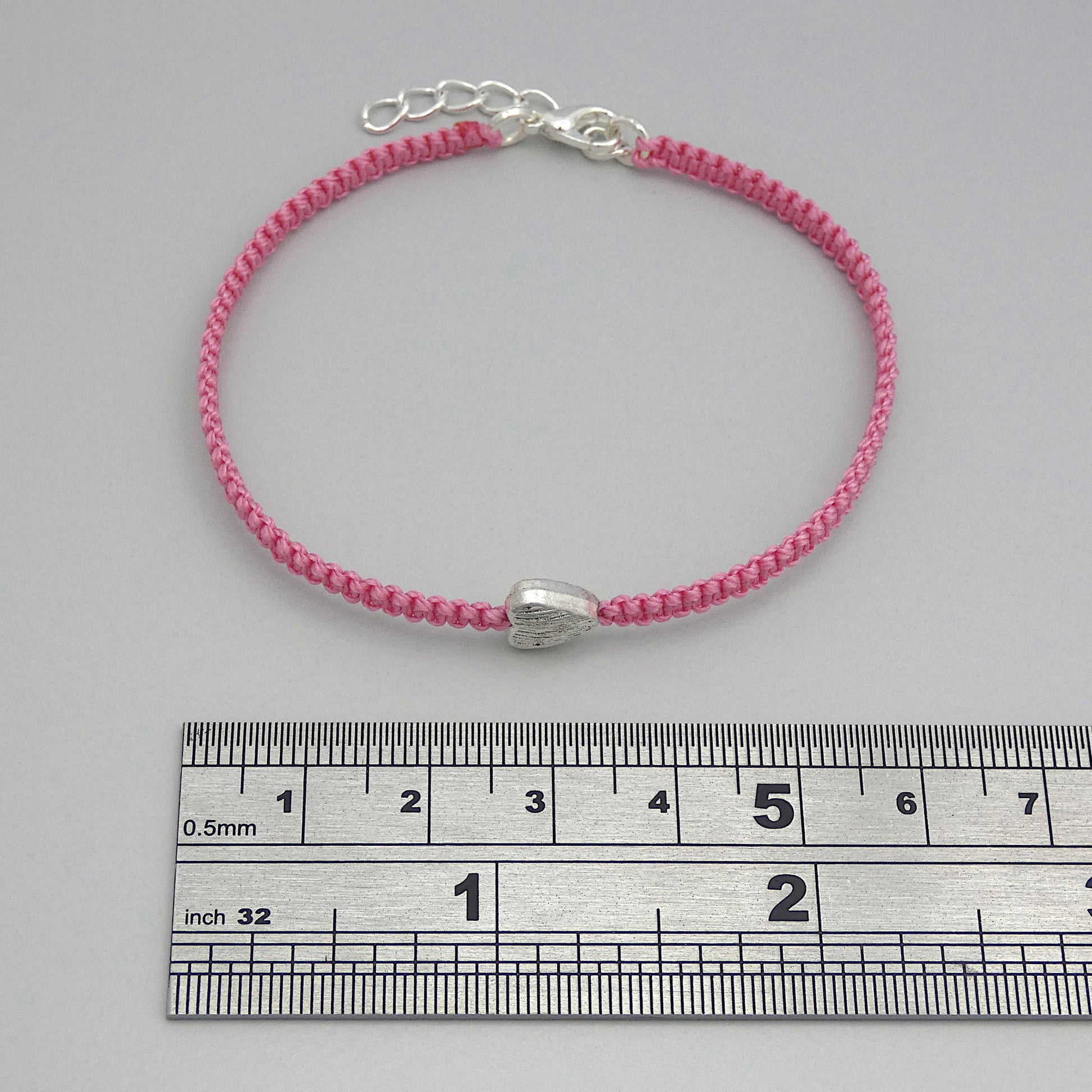 Pink seven inch macramé bracelet with silver plated textured heart charm, lobster clasp and one inch extender chain next to a ruler.